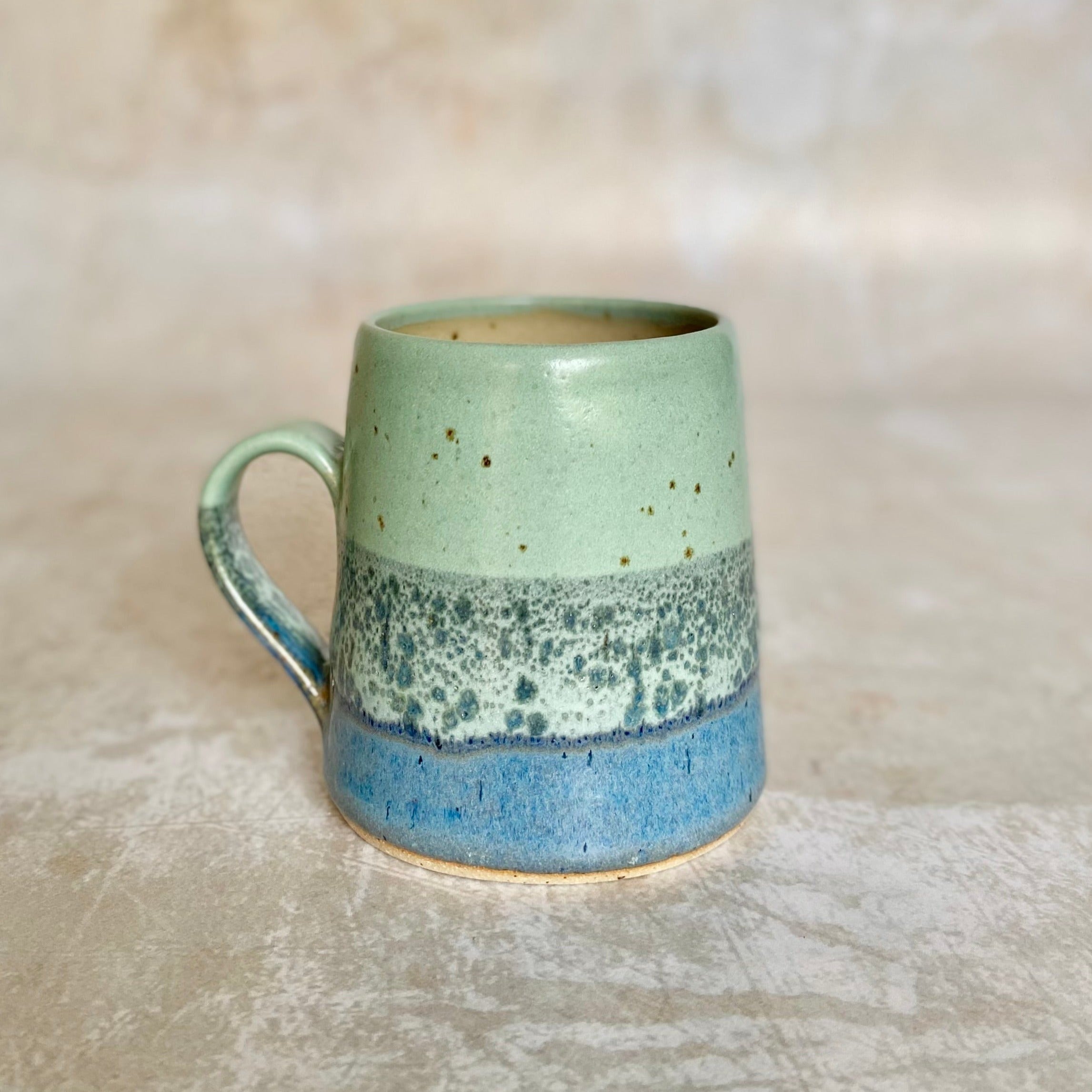 Hand-thrown ceramic mug with a light green body, speckled with brown, and a layered blue and white base.