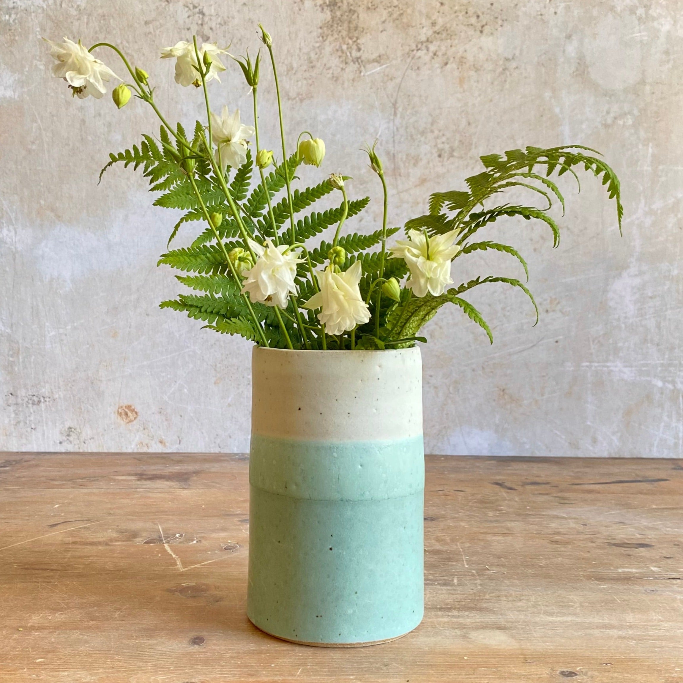 A speckled, cylindrical vase with a light cream top and a pale teal bottom holds a bouquet of white columbines and ferns.