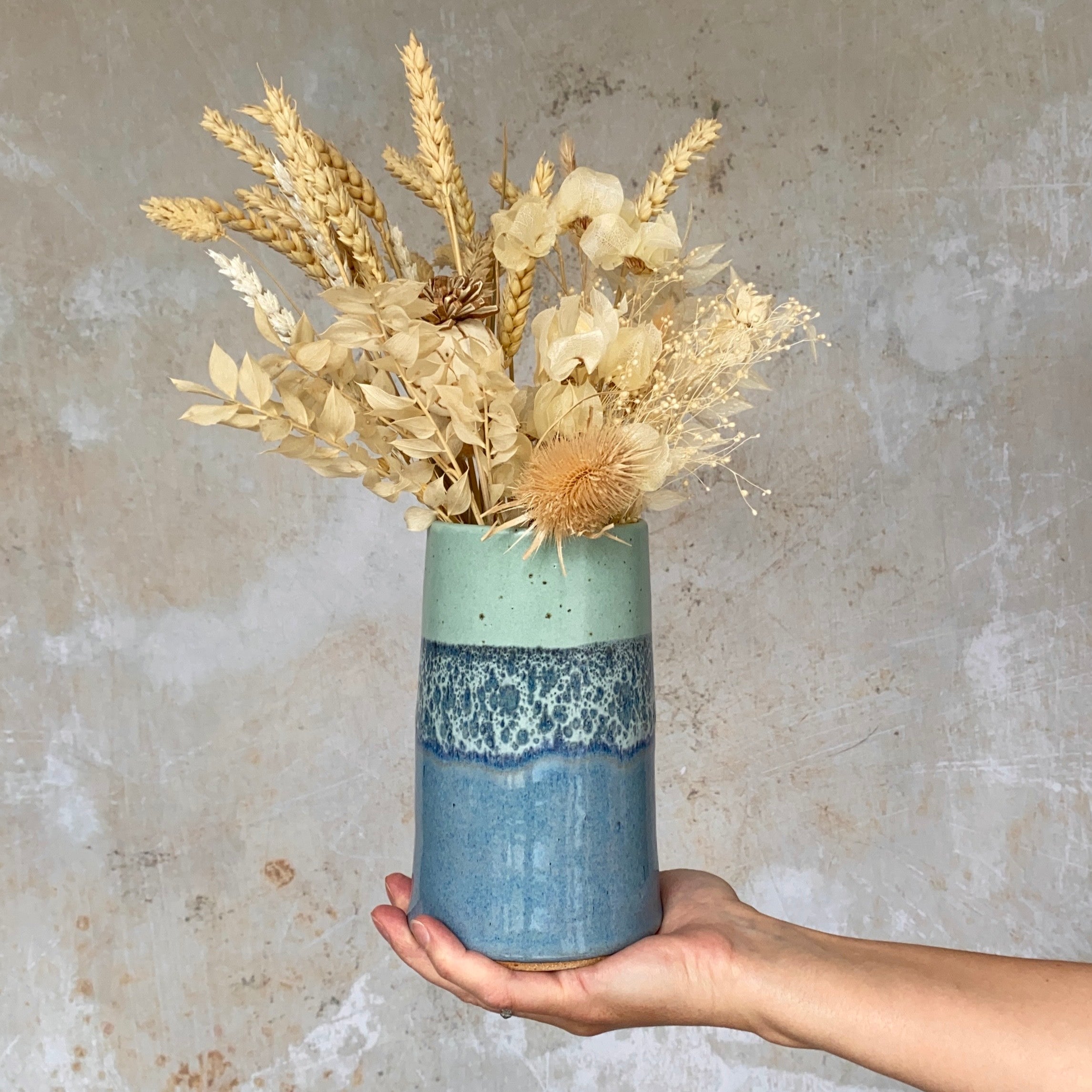 A blue and green speckled ceramic vase holds a bouquet of dried flowers, including wheat and thistle.