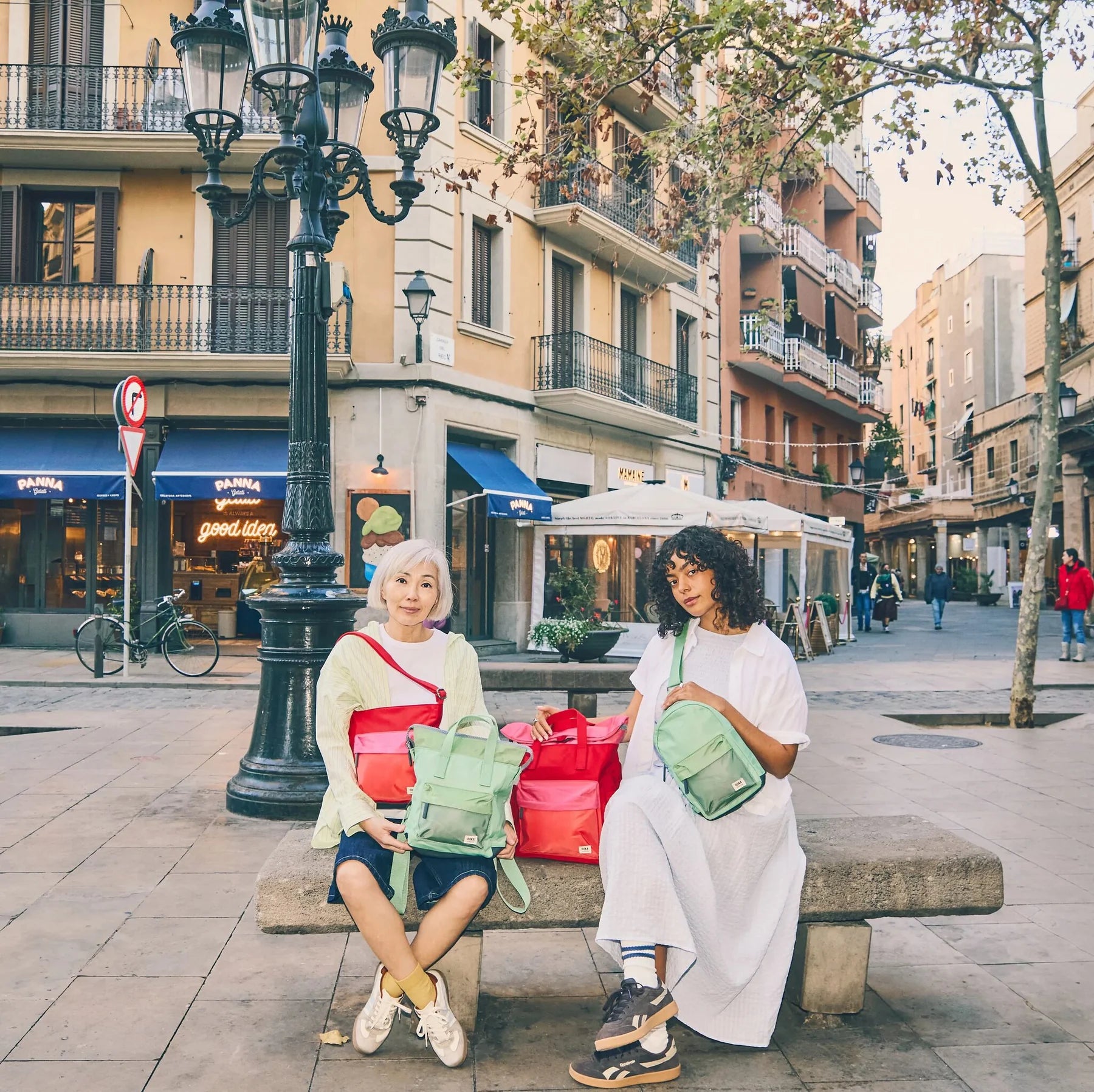 Two women sitting on a bench in an urban setting with colorful bags.