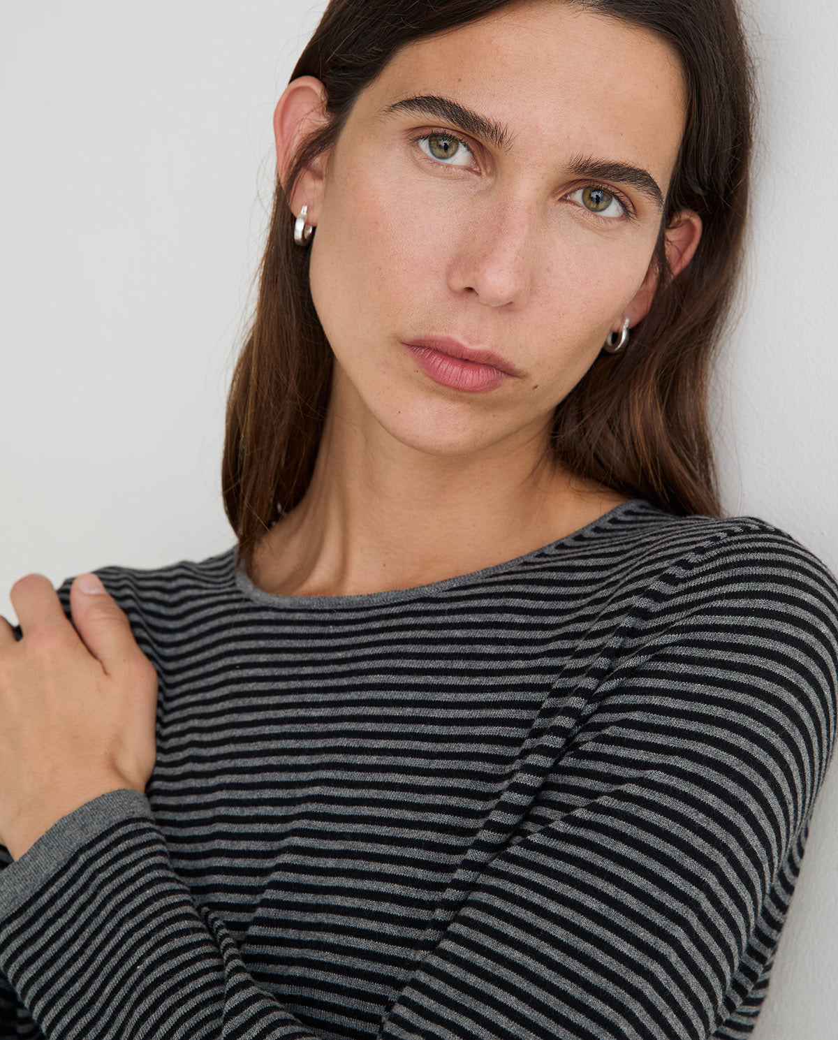 A close-up portrait of a woman wearing a dark gray and black striped long-sleeve top, with small silver hoop earrings, against a plain light background.
