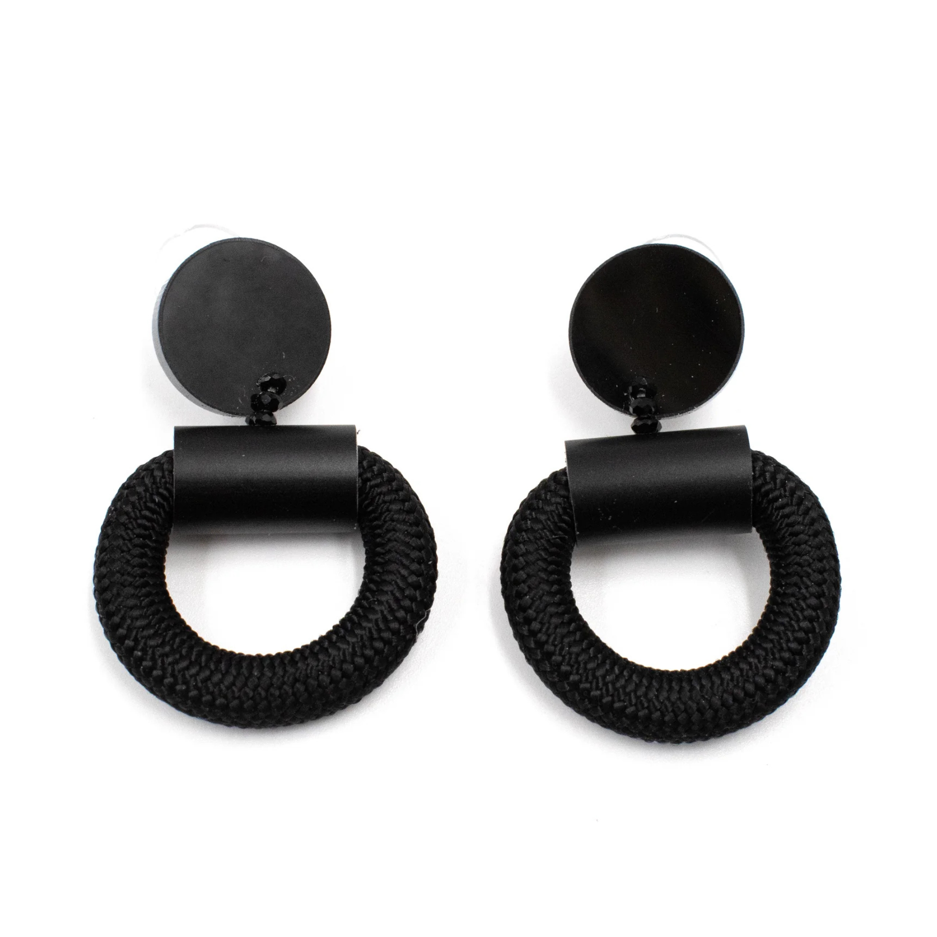 A pair of black statement earrings featuring a thick, braided circular ring suspended from a matte black cylindrical connector and a round disc, shown on a plain white background.