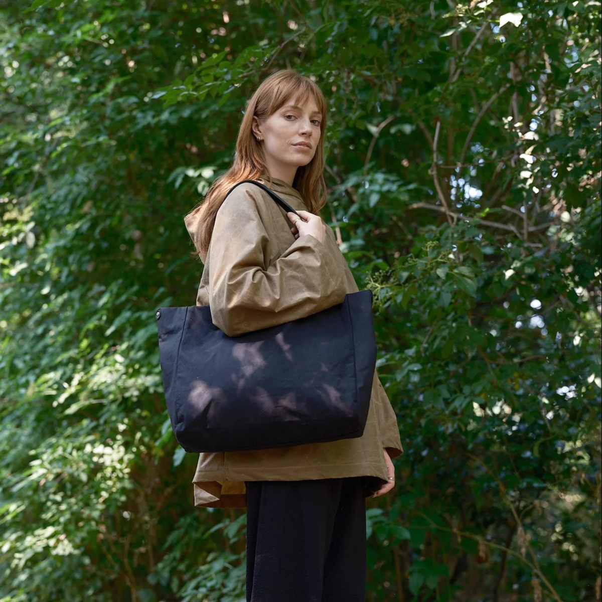 A woman wearing a tan jacket and black pants stands outdoors in a wooded area, carrying a large black tote bag over her shoulder.