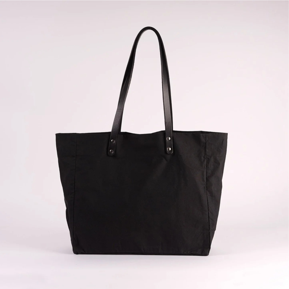 A black canvas tote bag with black shoulder straps and silver-tone metal rivets, photographed against a light gray studio background.