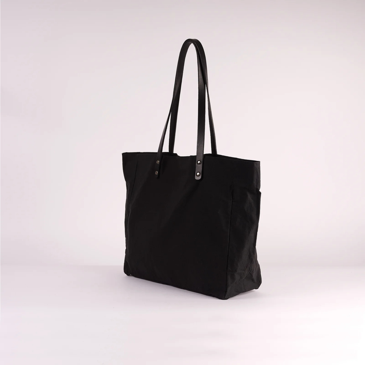 A black canvas tote bag with black shoulder straps and silver-tone metal rivets, photographed on a seamless light gray studio background.