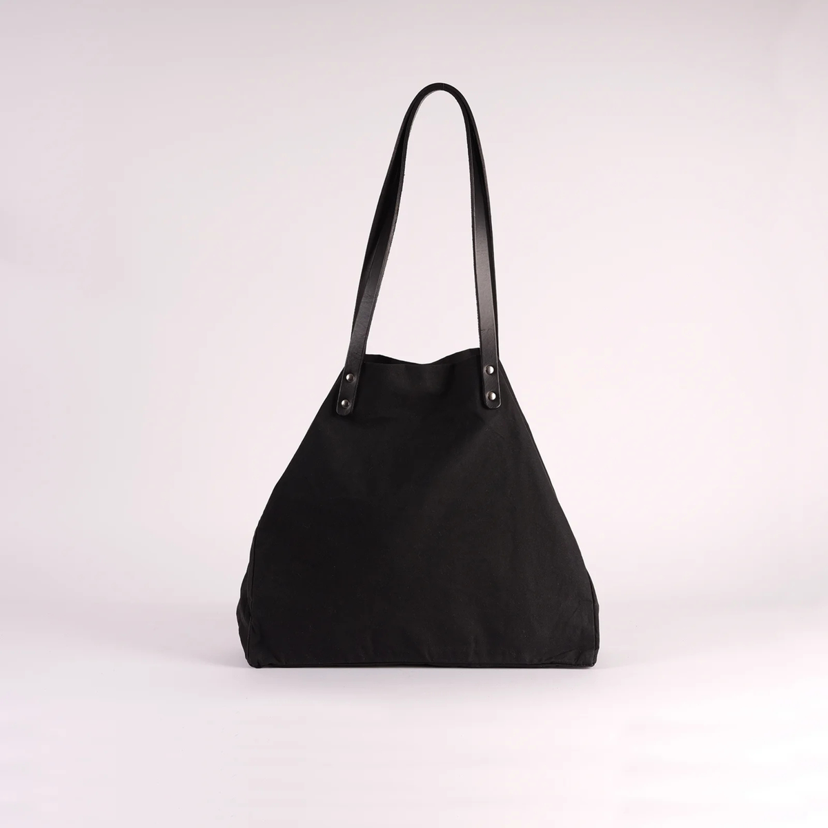 A black canvas shoulder bag with black leather straps and silver-tone metal rivets, photographed against a plain light gray studio background.
