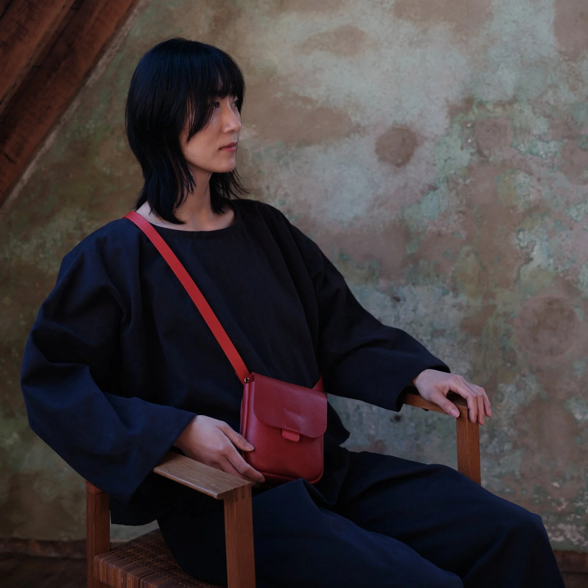 A woman with short black hair sits on a wooden chair against a textured plaster wall, wearing a black outfit and carrying a small red leather crossbody bag.