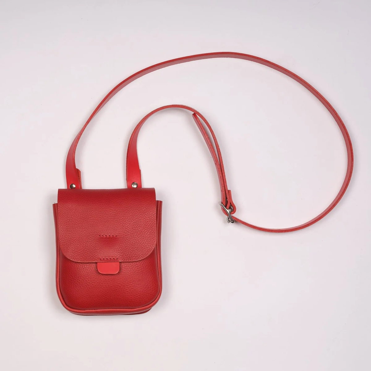 A small red pebbled leather crossbody bag with a long matching shoulder strap, laid flat on a light off-white background.