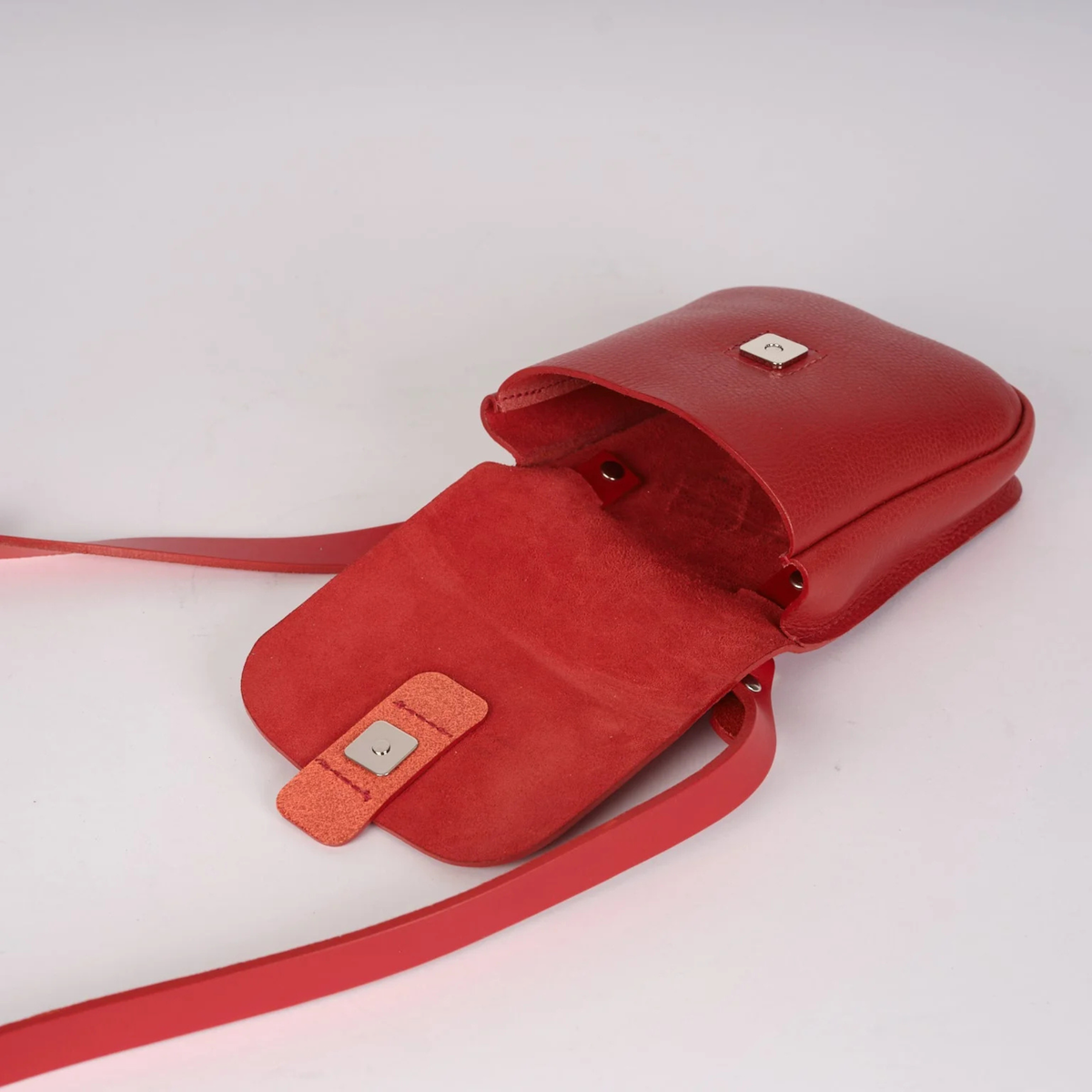 A red leather crossbody bag with an open flap and long strap, photographed on a clean white background. A small square metal logo plaque is visible on the flap and attached to the strap tab.