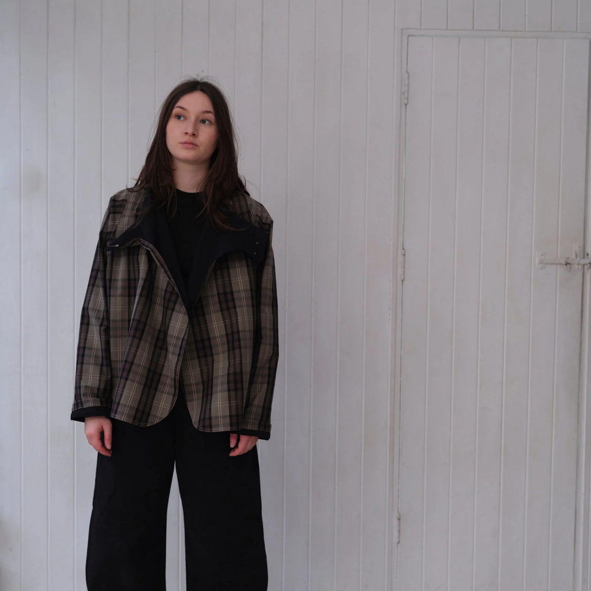 A woman stands against a white paneled wall wearing a brown and black plaid jacket over a black top and black wide-leg pants.