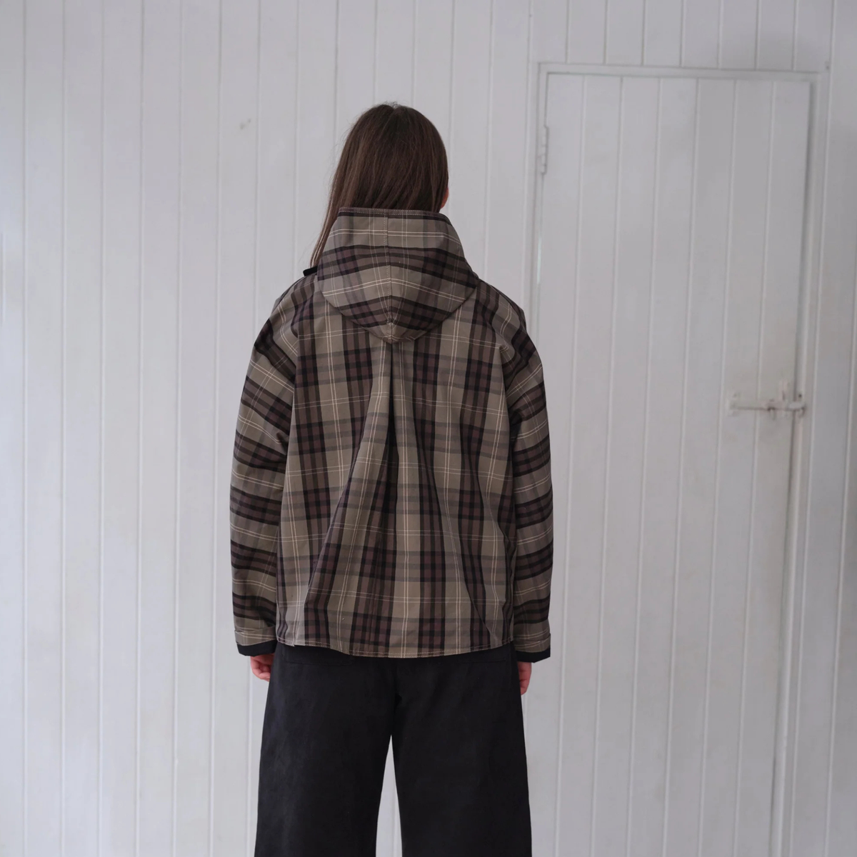 A person viewed from behind wearing a brown and beige plaid hooded jacket and dark pants, standing in front of a white paneled wall and door.