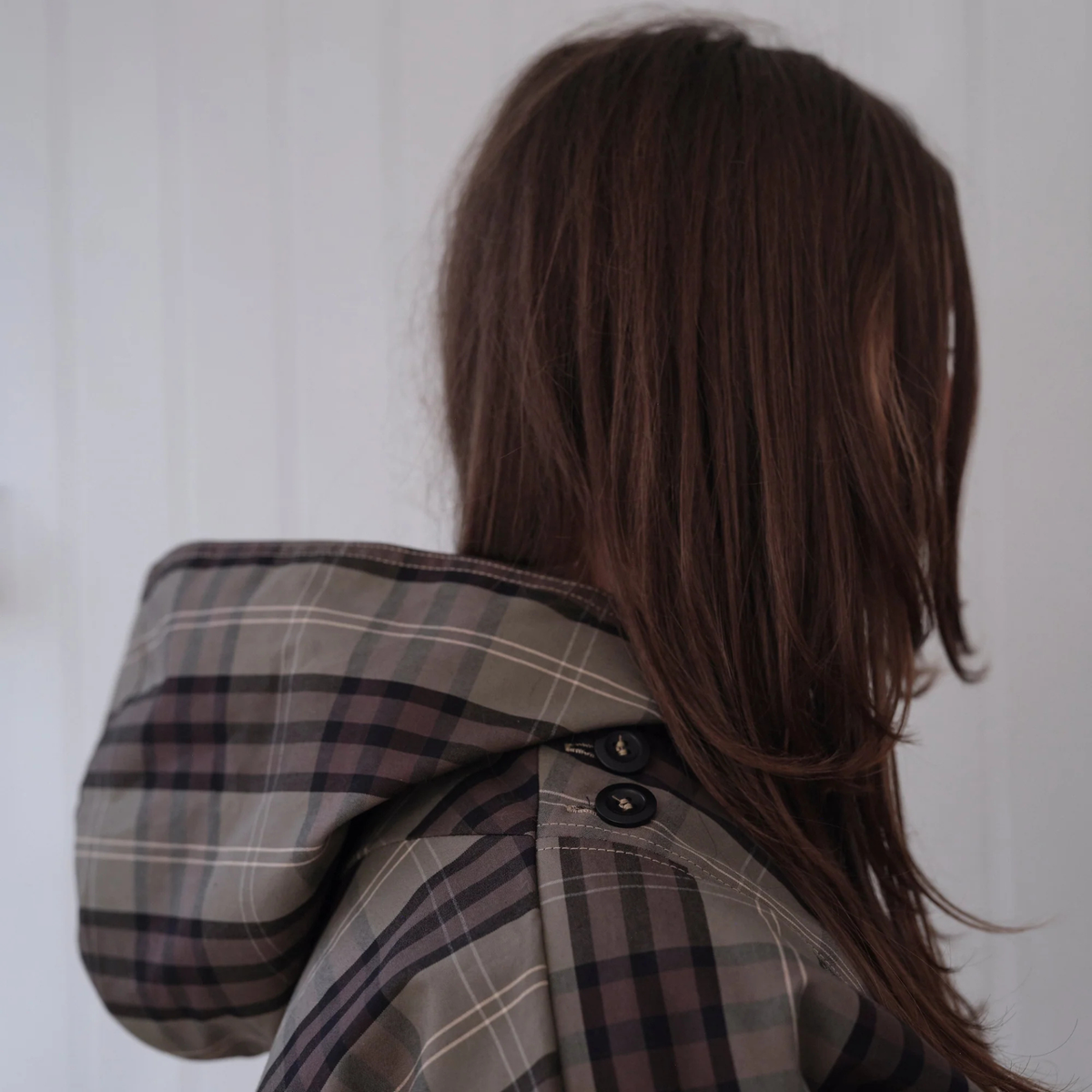 A person shown from the back wearing a plaid hooded jacket in muted gray, brown, and black tones, with long brown hair visible against a soft light background.