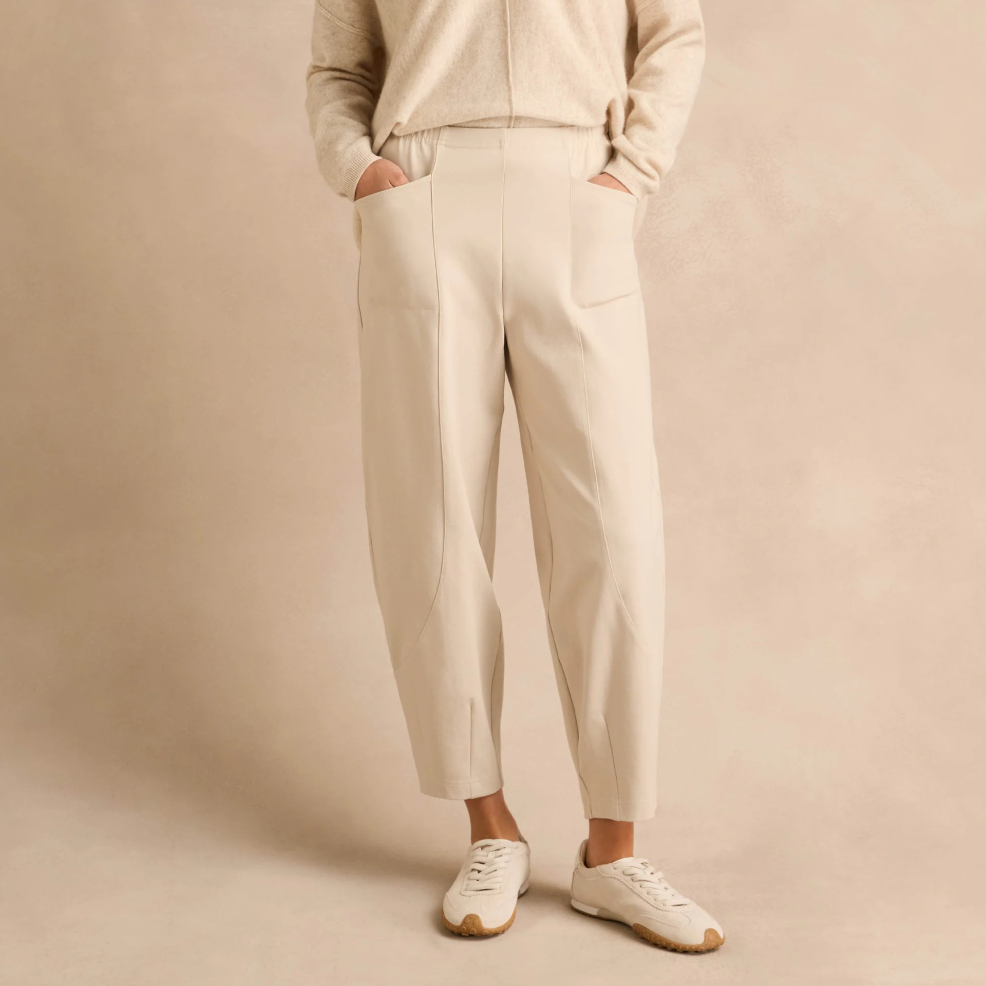 A person wearing cream-colored pleated trousers with a matching knit top and white sneakers, standing against a warm beige studio backdrop.