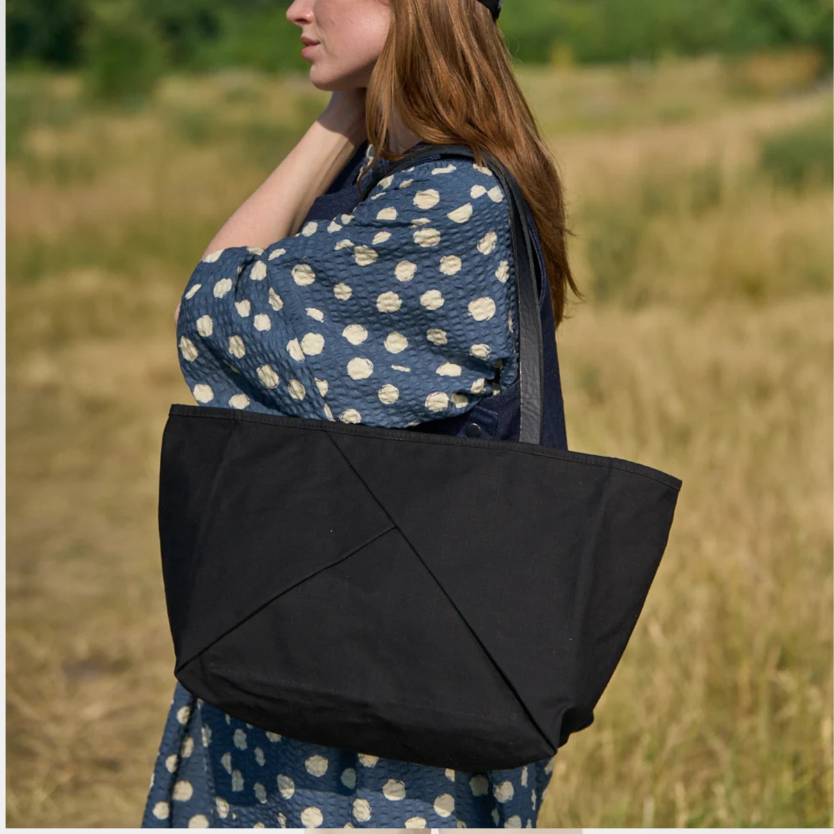 A woman in a blue and white polka dot dress carries a large black canvas tote bag over her shoulder in an outdoor grassy field.