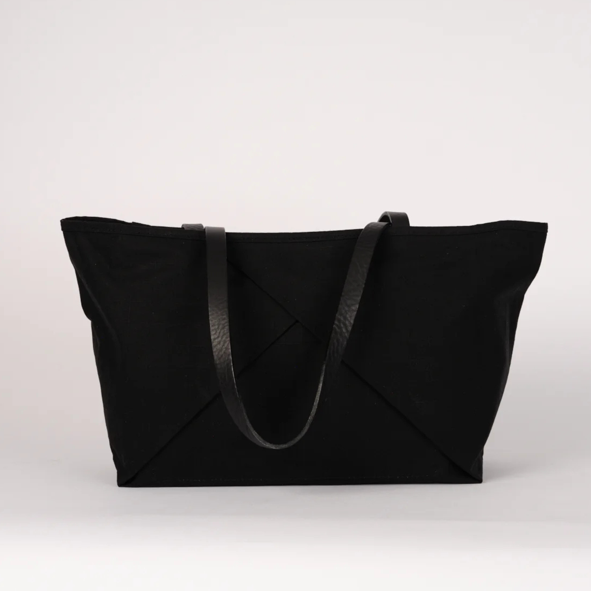 A black canvas tote bag with black leather shoulder straps, displayed against a clean, light gray studio background.