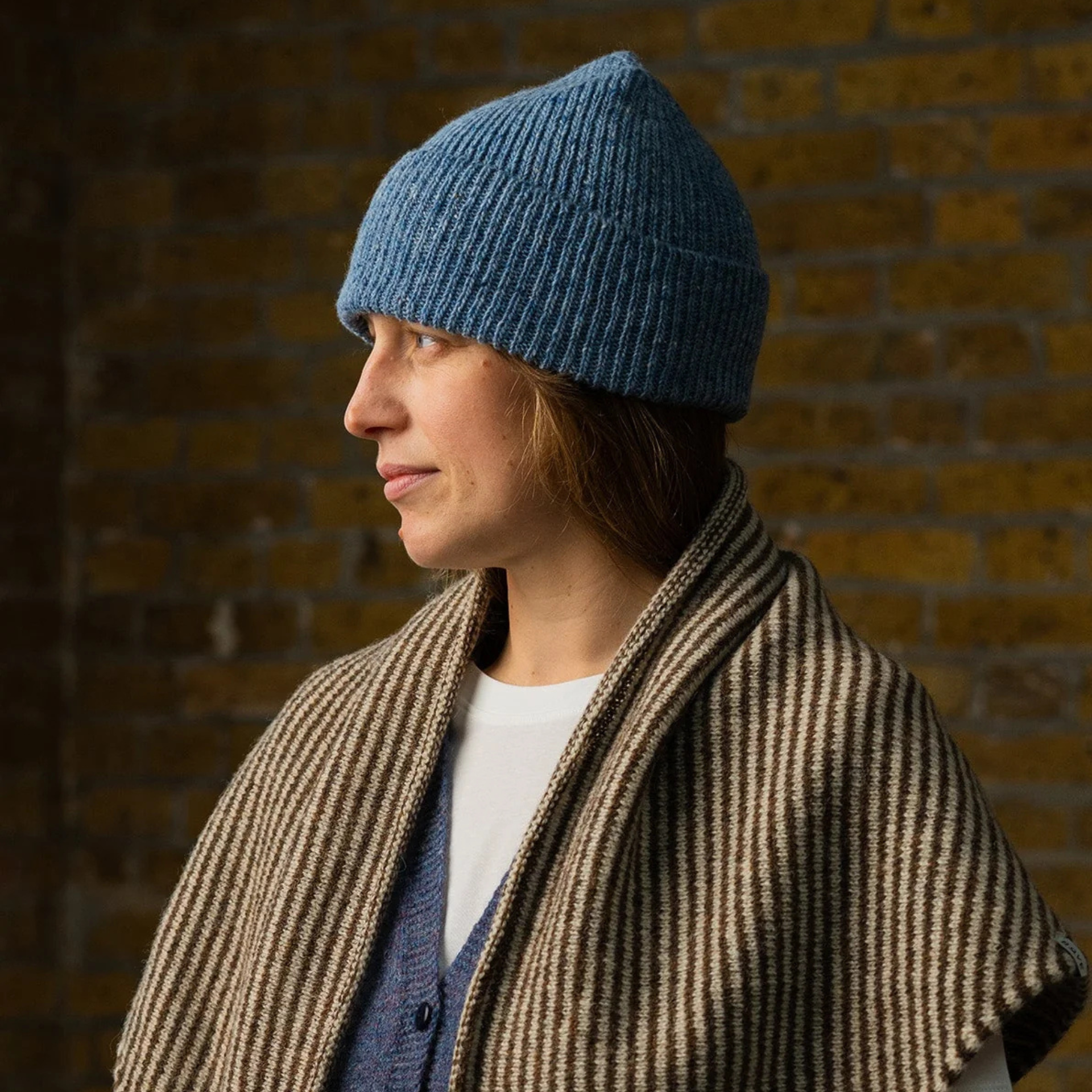 A woman in profile wearing a blue ribbed knit beanie and a brown and beige striped shawl over a white shirt and blue cardigan, set against a warm brick wall background.