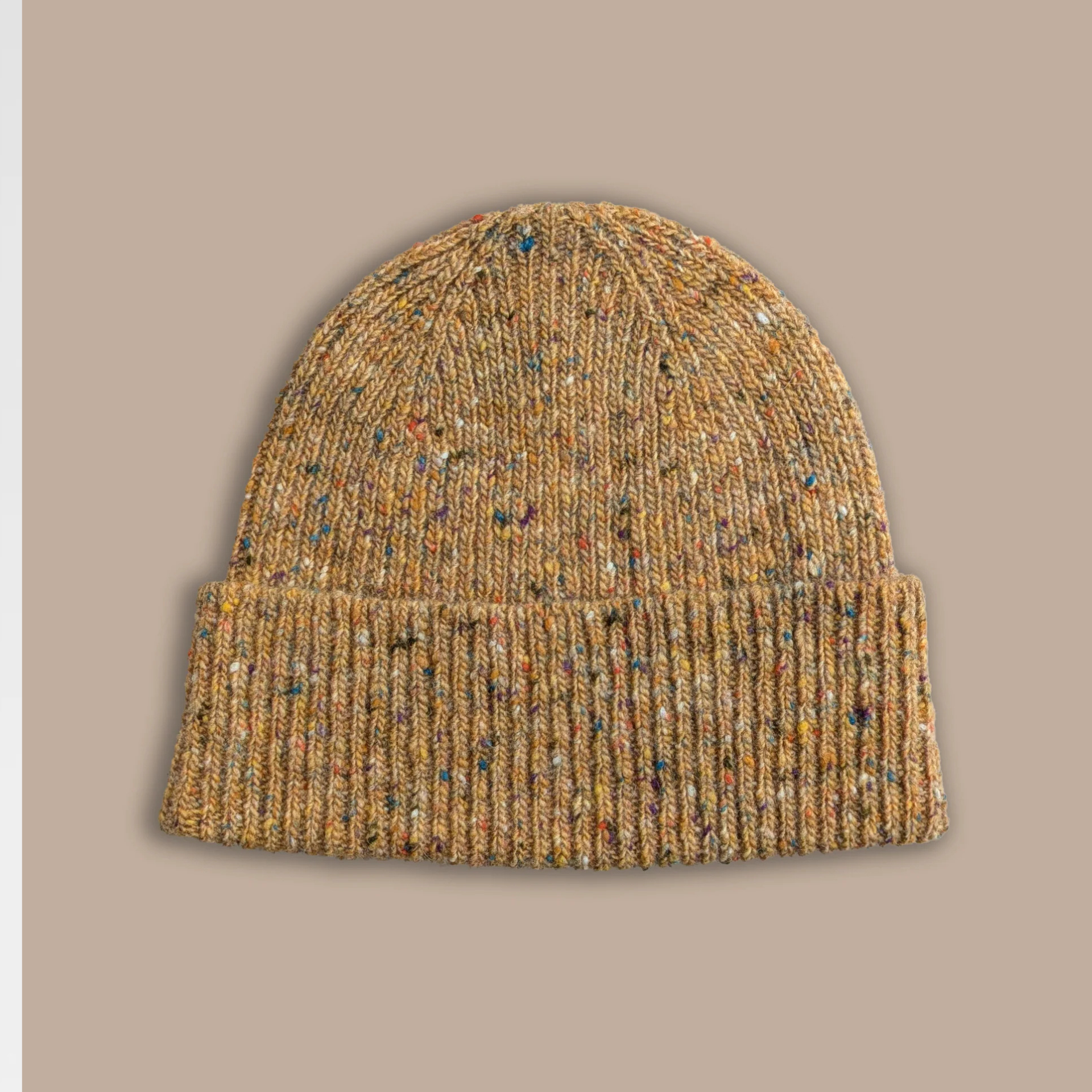 A ribbed knit beanie hat in a warm tan and brown heathered yarn with small multicolored flecks, shown against a solid taupe background.