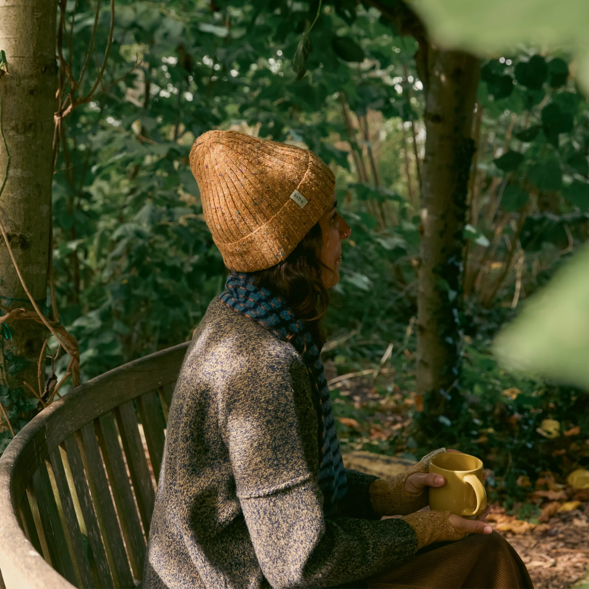 A woman sits on a wooden bench in a wooded outdoor setting, wearing a ribbed tan knit beanie, a textured taupe cardigan, and a blue patterned scarf while holding a yellow mug.