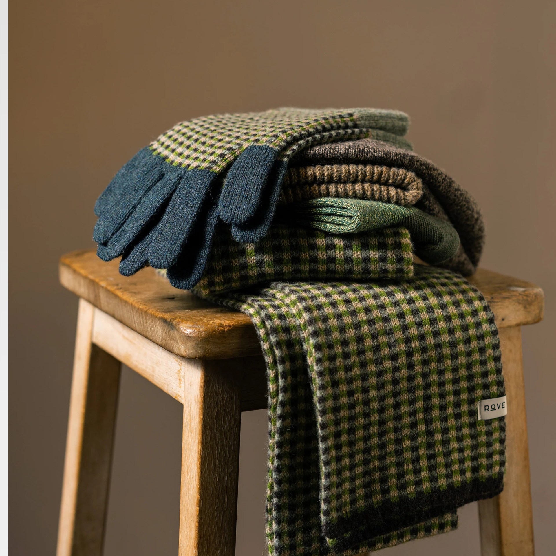 A stack of folded knit winter accessories, including gloves and a scarf, displayed on a wooden stool against a warm brown background. The items feature a green and black houndstooth pattern with solid dark blue-green glove palms and cuffs.