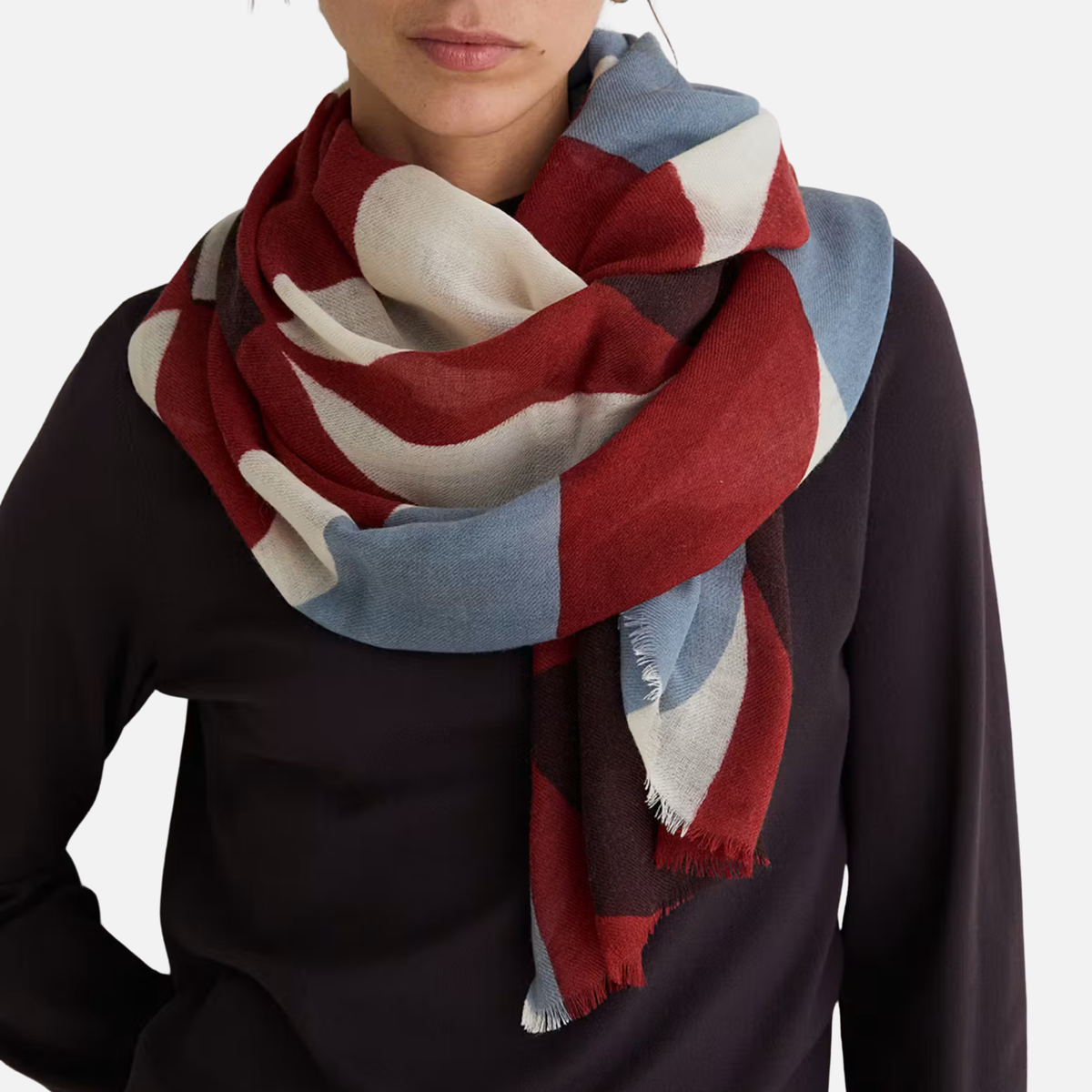 A close-up of a person wearing a large multicolor scarf with bold blocks of red, cream, light blue, and dark brown over a black top, against a clean white background.