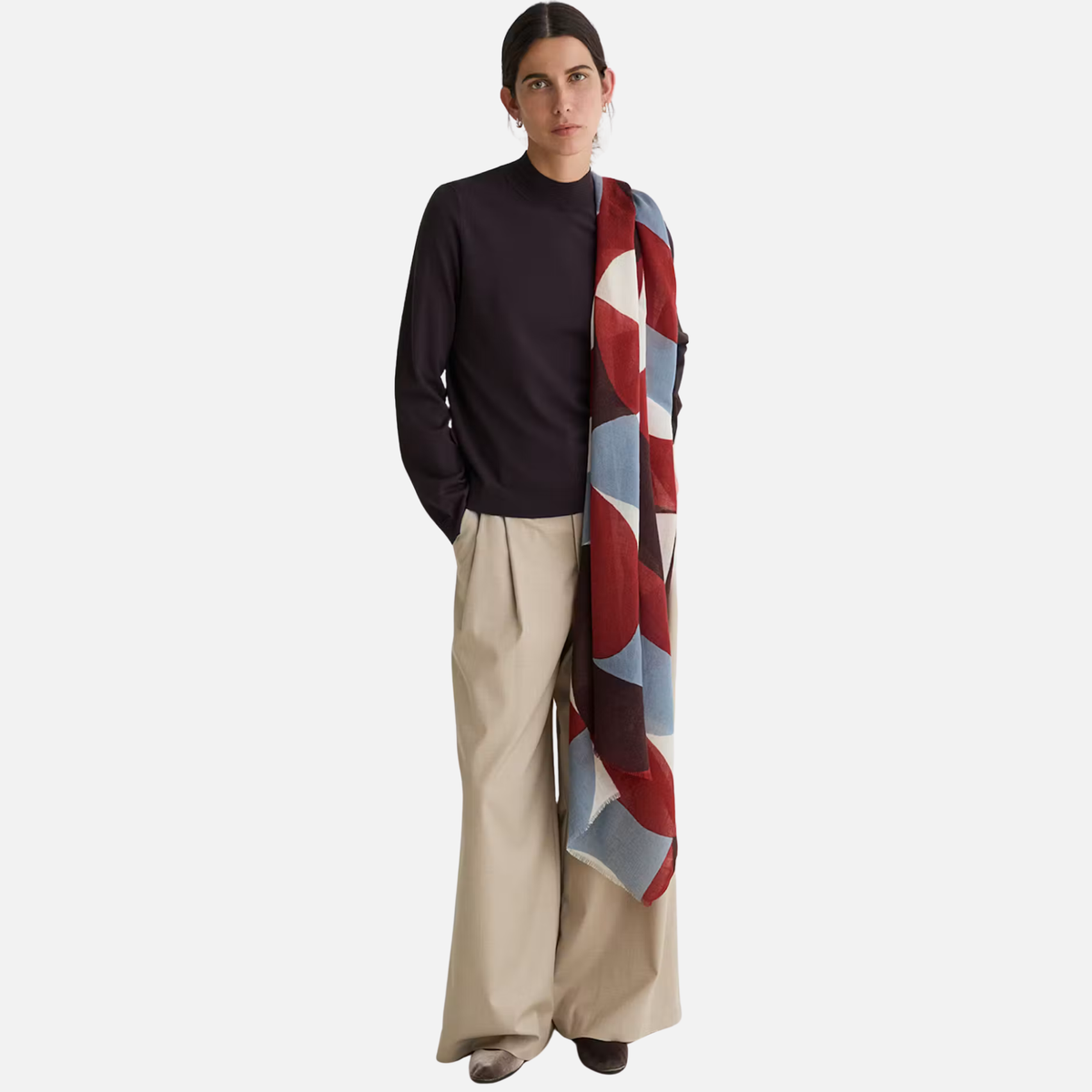 A model stands against a light gray studio background wearing a dark navy mock-neck long-sleeve top, beige wide-leg trousers, and a long patterned scarf with red, blue, and cream geometric shapes.