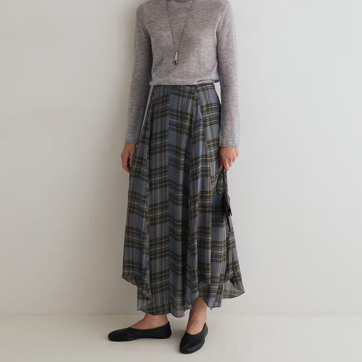A model wearing a gray knit long-sleeve top and a gray plaid maxi skirt with black flats, standing against a plain off-white wall.