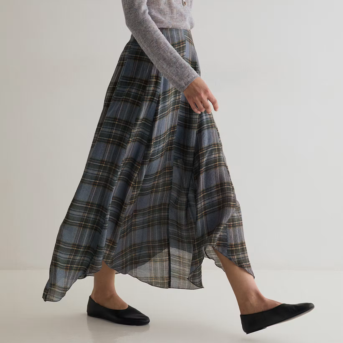 A person wearing a long plaid midi skirt in muted blue, gray, and brown tones, paired with a light gray long-sleeve top and black flats, shown against a plain light gray studio background.