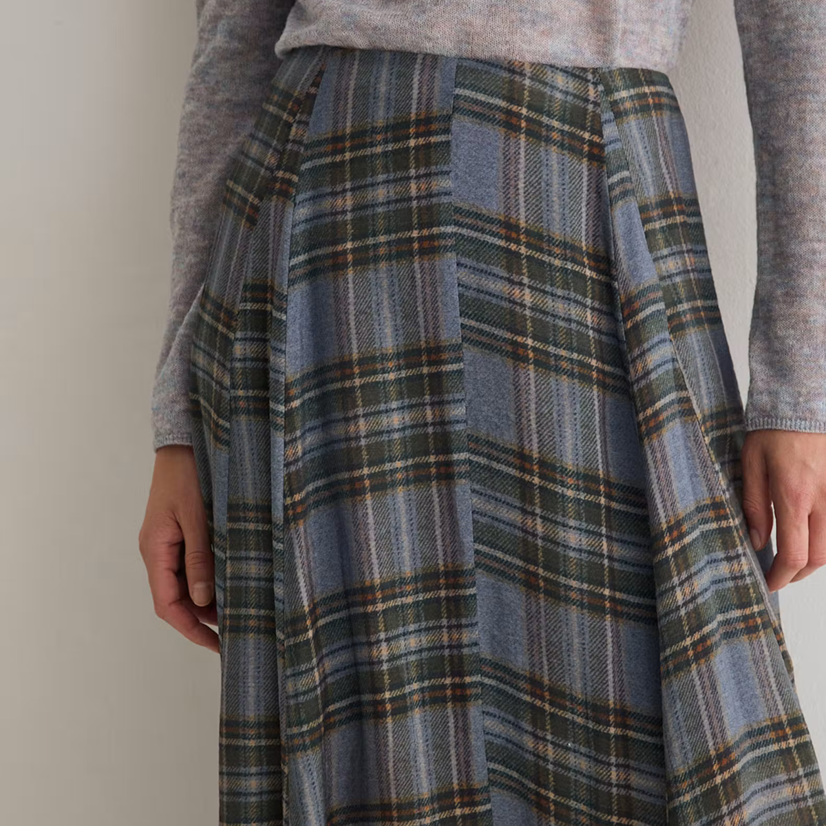 A close-up of a person wearing a high-waisted, pleated plaid skirt in muted blue, green, and beige tones, paired with a gray knit top against a plain light background.
