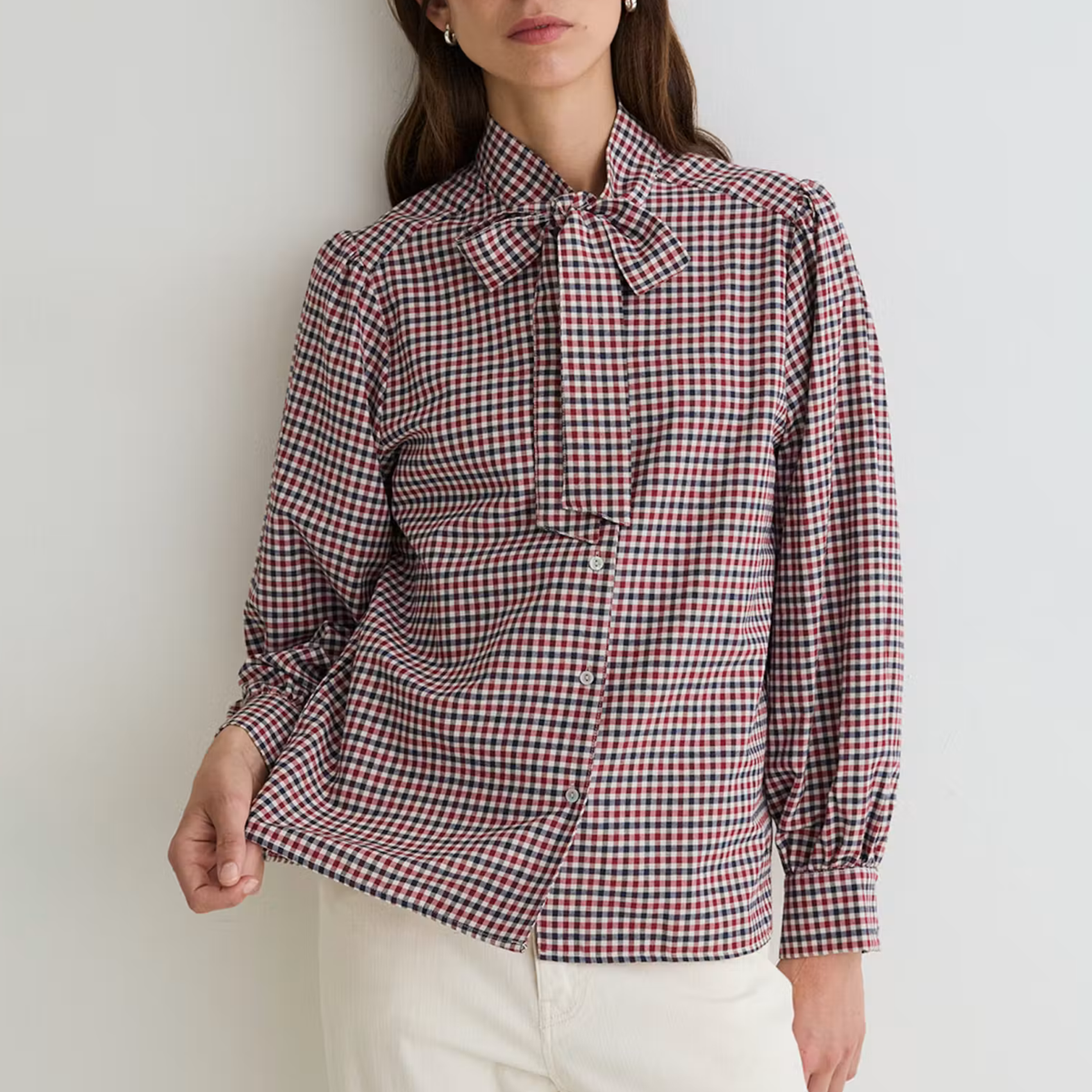 A woman wearing a red, white, and blue gingham long-sleeve blouse with a bow tie collar, paired with off-white trousers, standing against a plain light gray wall.