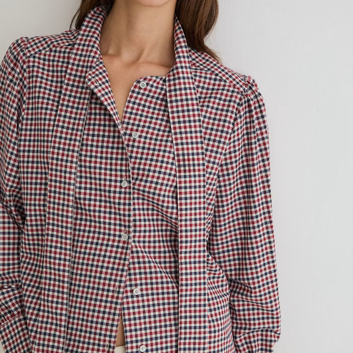 A close-up of a model wearing a red, black, and white gingham check long-sleeve button-up shirt. The shirt features a relaxed fit with a wide collar and buttoned front, shown against a plain light gray background.