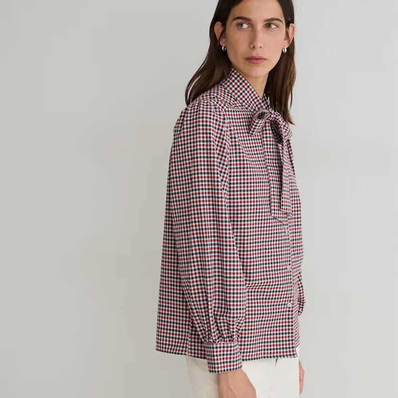 A woman wearing a red, white, and blue gingham long-sleeve blouse with a tie-neck detail, shown from a side profile against a light gray studio background.