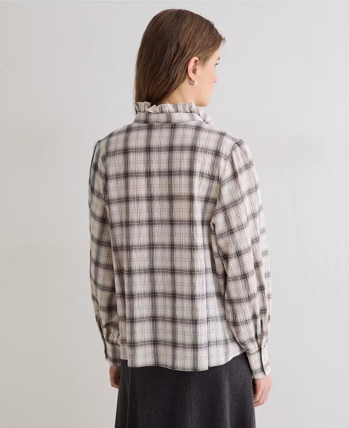A woman shown from the back wearing a long-sleeve plaid shirt with a ruffled high collar, in a light beige and dark gray check pattern, paired with a dark skirt against a plain light gray studio background.