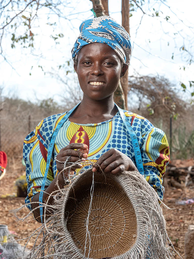Woman weaving a large, round basket with light gray fibers; the basket has a textured, circular pattern.