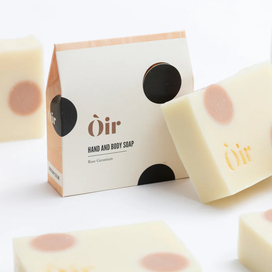 Bar soap with packaging that reads 'Oir Hand & Body Soap' Rose Geranium Scent. The packaging has a minimalist design with black dots and a hint of color on the lower right corner.