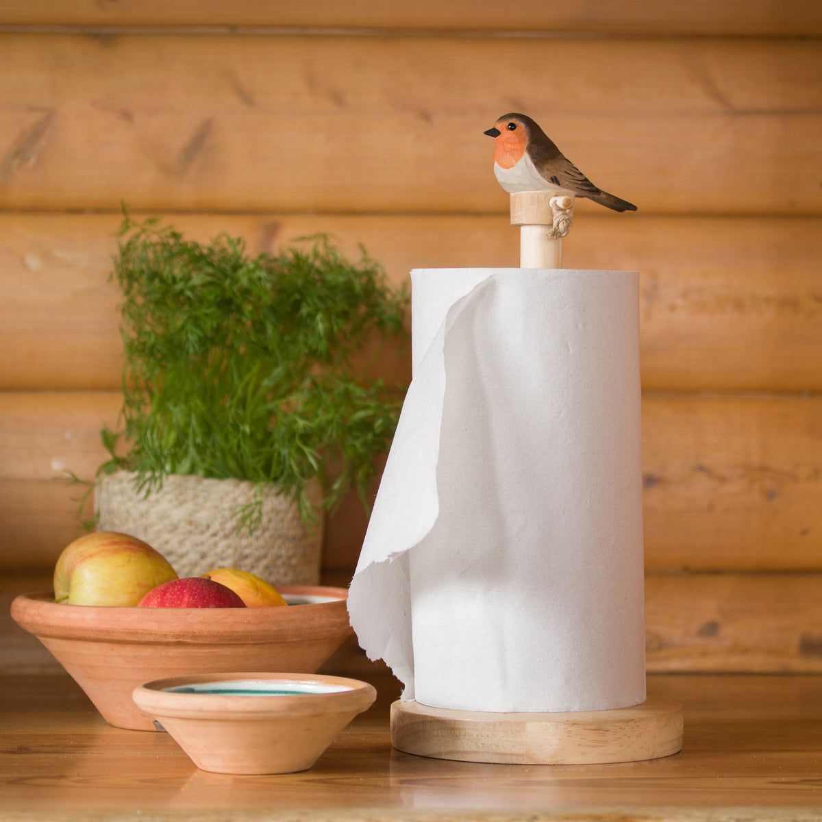Paper towel holder with a decorative bird on top, placed on a wooden surface with a plant and fruit bowl in the background.
