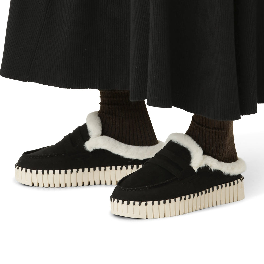Black suede loafers with cream faux fur trim and a cream and black textured sole. The loafers are worn with dark brown socks and