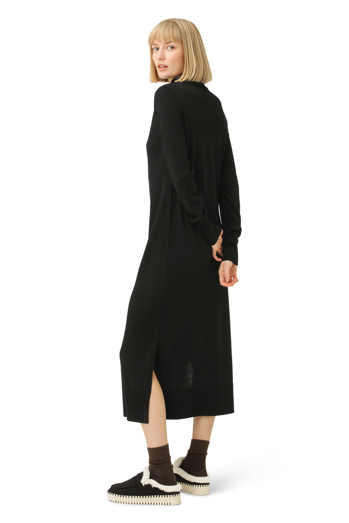 Woman in black midi dress, back view, long sleeves, side slits, wearing brown socks and dark brown shearling-lined slippers.