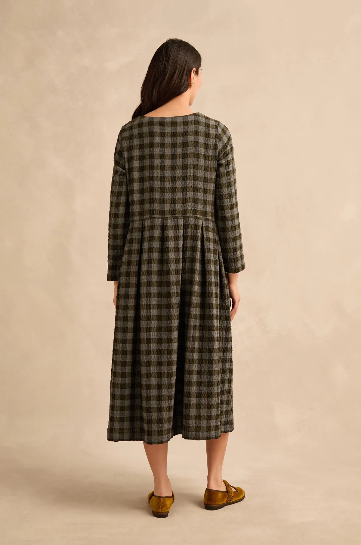 A woman viewed from behind wearing a long-sleeved, knee-length plaid dress in olive green and black, paired with mustard yellow slip-on shoes, against a textured beige studio backdrop.