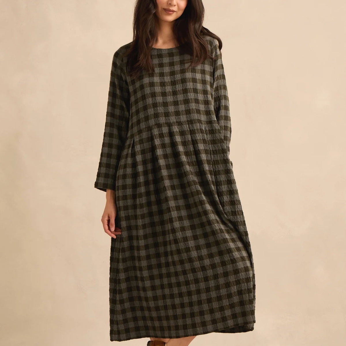 A woman modeling a long-sleeved, knee-length plaid dress in dark olive green and black, paired with brown suede sandals, against a solid beige studio backdrop.