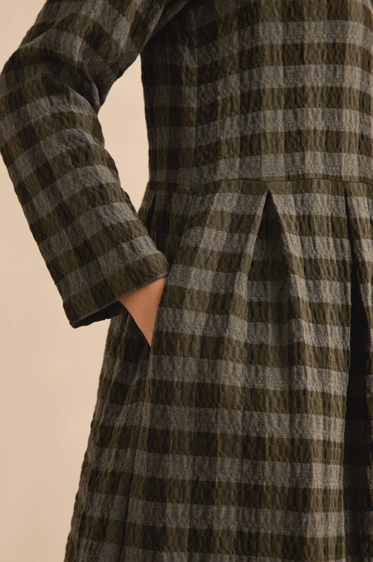 Close-up of a person wearing a textured plaid coat or dress in olive green and gray, with one hand in a pocket against a plain beige background.