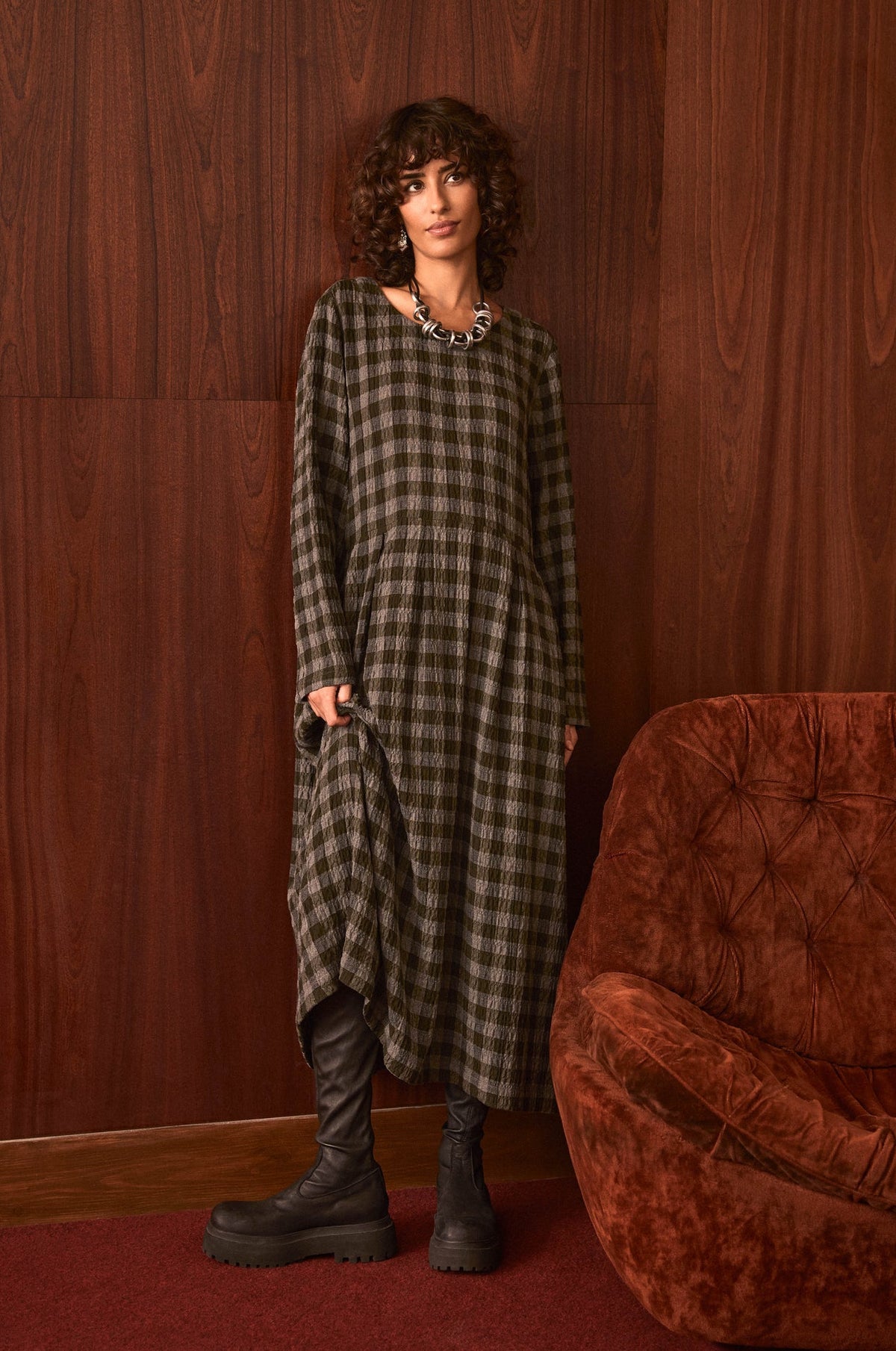 A model stands in a room with dark wood paneling and a rust-colored armchair, wearing a long-sleeved olive green and black plaid dress with black boots.