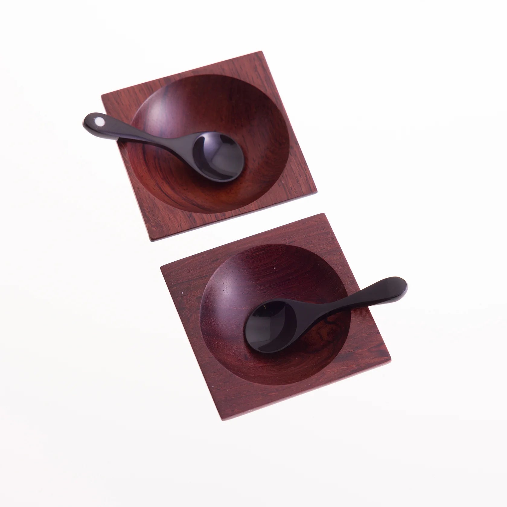 Two dark-red square wooden bowls, each containing a small black spoon.