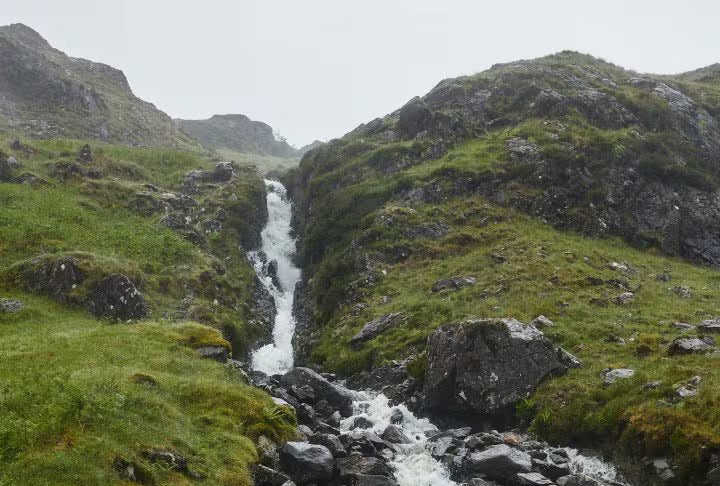 A rocky mountain stream with a small waterfall cascading down grassy hills under an overcast sky.