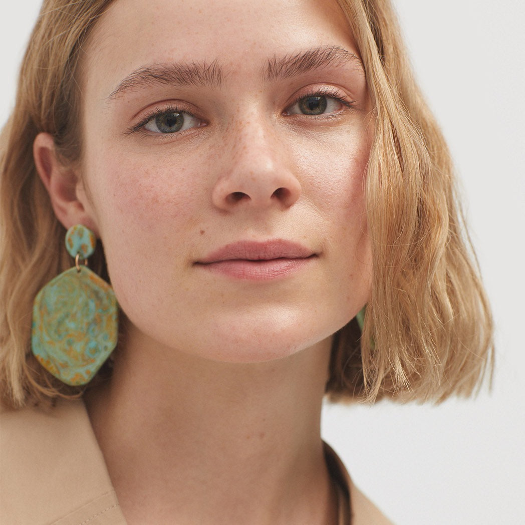 Woman wearing teal and gold patterned drop earrings.