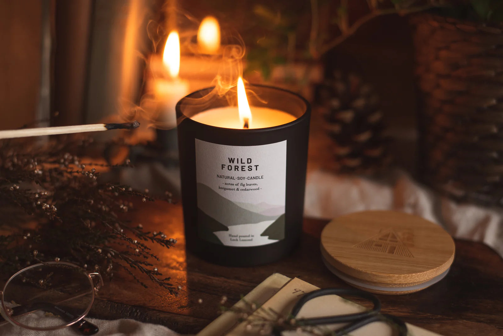 Lit black Wild Forest natural soy candle with fig leaf, bergamot, and cedarwood scent.  Bamboo lid visible.