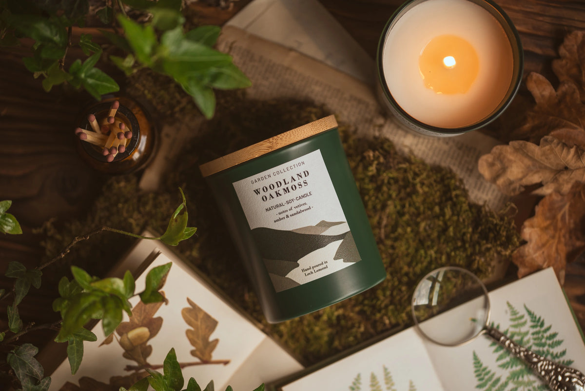 A dark green candle tin labeled 'Woodland Oakmoss' is styled in a cozy flat lay with a lit white candle, moss, leaves, a magnifying glass, and botanical cards on a rustic wooden surface.
