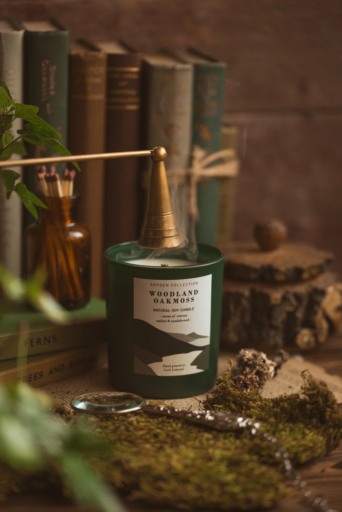 A dark green scented candle in a round tin with a brass wick and a white label reading 'Woodland Warmth' by Trader Collection, styled on a rustic surface with moss, books, and a matchbox in the background.