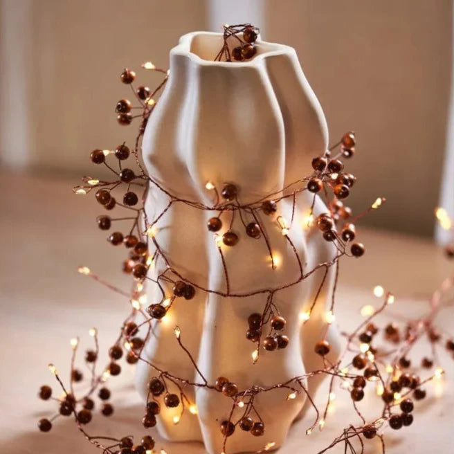 Copper wire string lights with warm white LEDs and brown berry accents wrapped around a decorative vase.