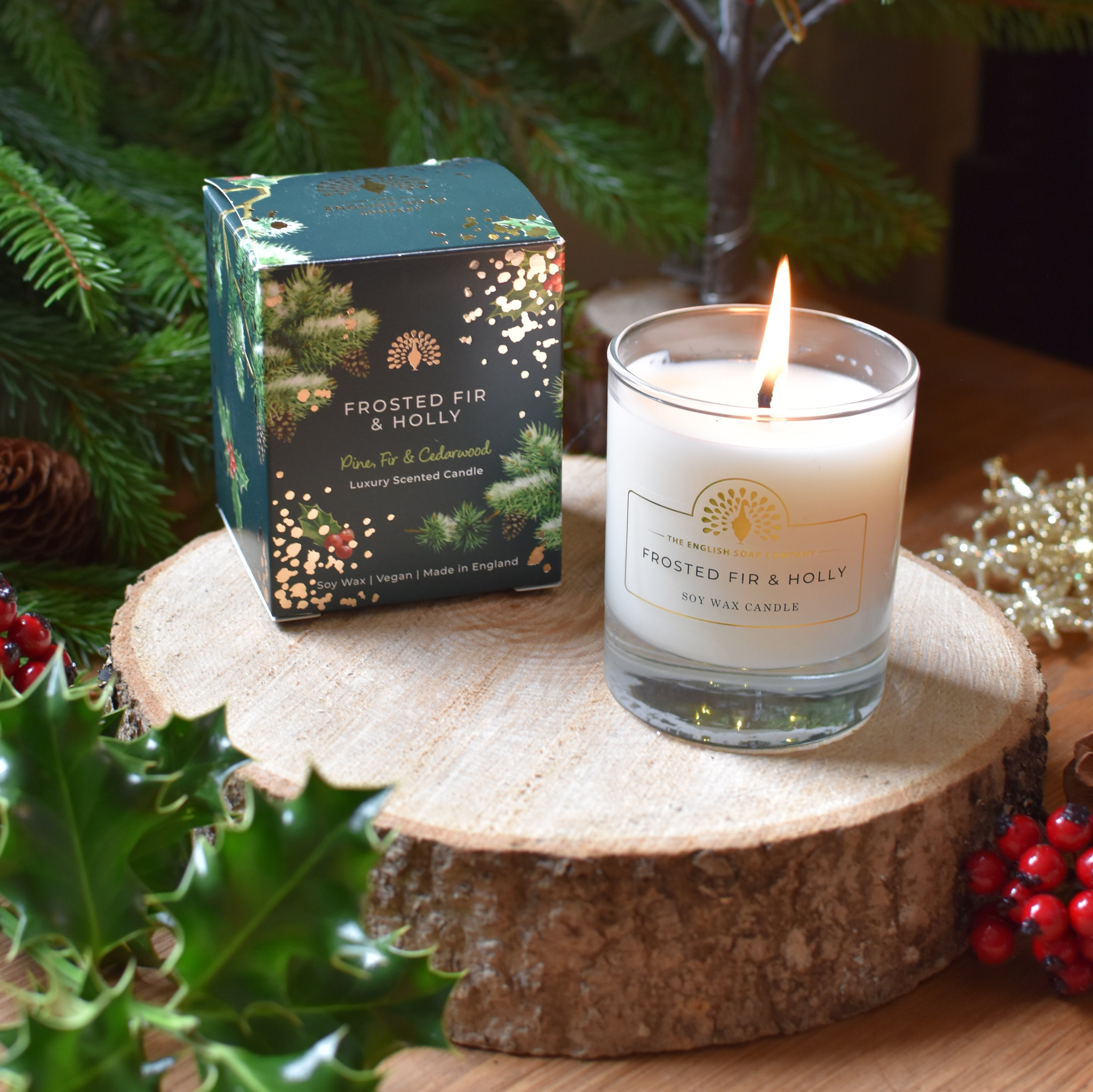 Frosted Fir & Holly soy wax candle burning in a clear glass jar, next to its dark green and gold packaging.