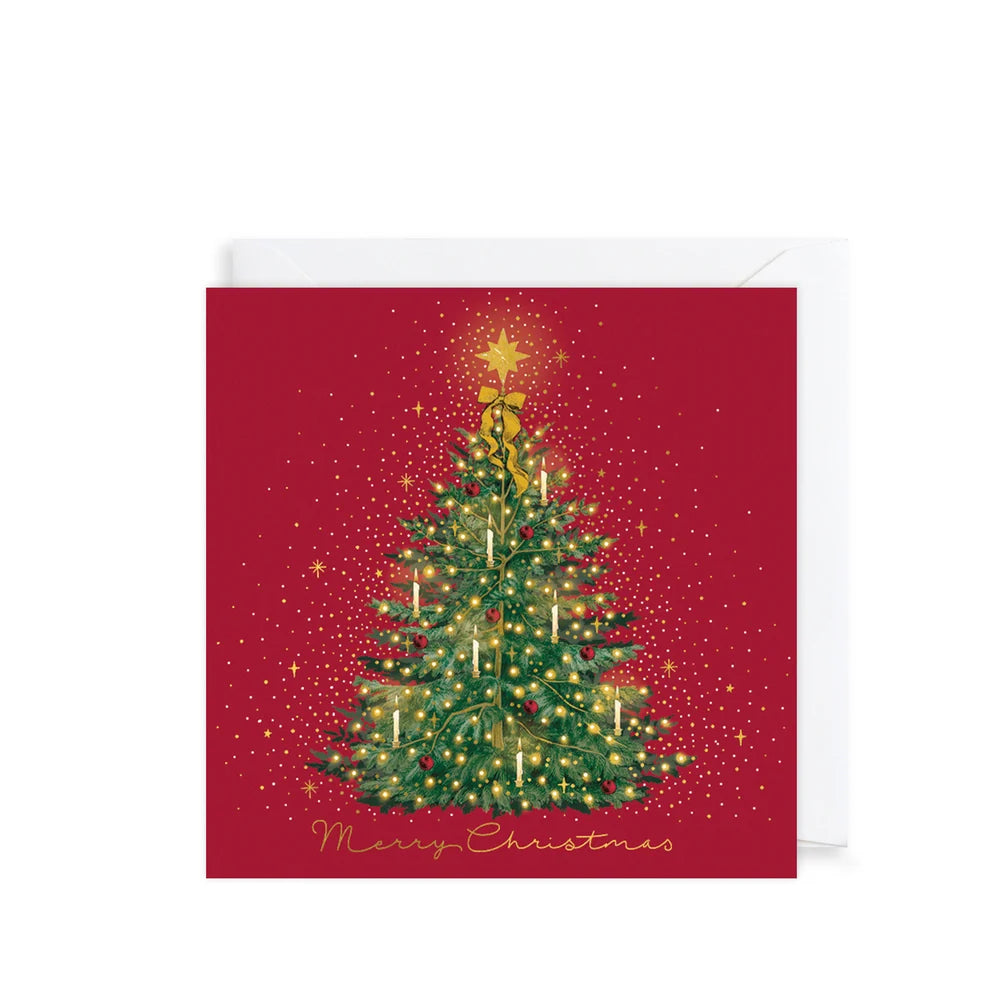A square Christmas greeting card with a red background, featuring an illustrated green Christmas tree with gold lights and a gold star, with 'Merry Christmas' written in gold script at the bottom. A white envelope is partially visible behind the card.